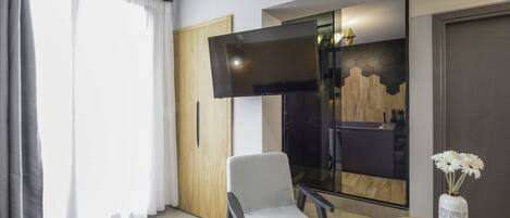 A modern hotel room with a large flat-screen TV, a grey armchair, a wooden door, and a vase with flowers on a round table.