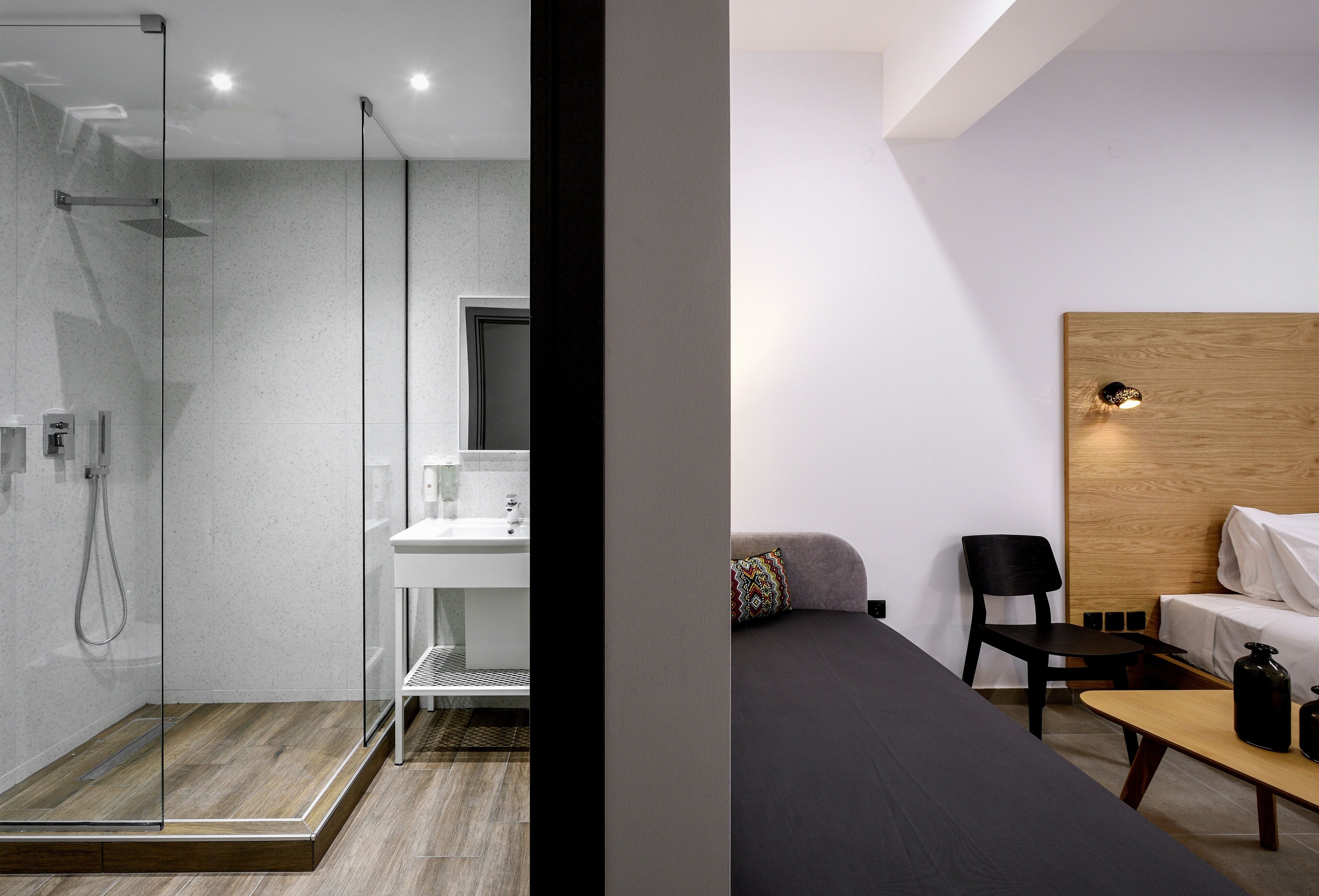 Habitación de hotel moderna con ducha de vidrio, suelo de madera, lavamanos blanco, cama con cabecera negra y una mesita con una botella.