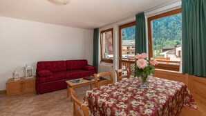 A cozy room with a red sofa, a wooden table with a floral tablecloth, and a view of a mountain through large windows. - Apartment in the heart of Valle di Sole (Mezzana-Marilleva)