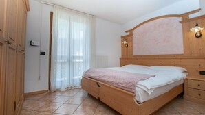 A bedroom with a wooden bed, a nightstand, a dresser, a radiator, and a window with sheer curtains. - Apartment in the heart of Valle di Sole (Mezzana-Marilleva)