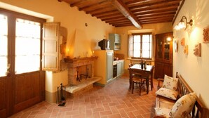 A rustic interior with a fireplace, wooden beams, a dining area with a table and chairs, and a kitchen with cabinets and a refrigerator. - Podere San Gregorio (Pienza)
