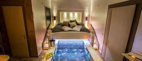 A modern hotel room with a large bed, a built-in jacuzzi, and a small table with a lamp.