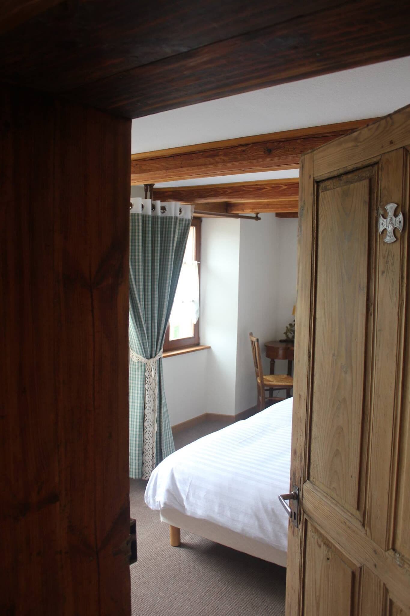 Une pièce avec une porte en bois, un lit avec du linge de lit blanc, une fenêtre avec un rideau et une chaise.