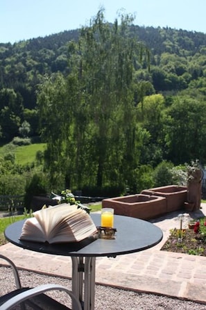 An outdoor table with a book and a glass of orange juice, surrounded by a garden and a view of a forested hill. - Chambres d'Hotes A l'Ombre du Verger (Breitenau)