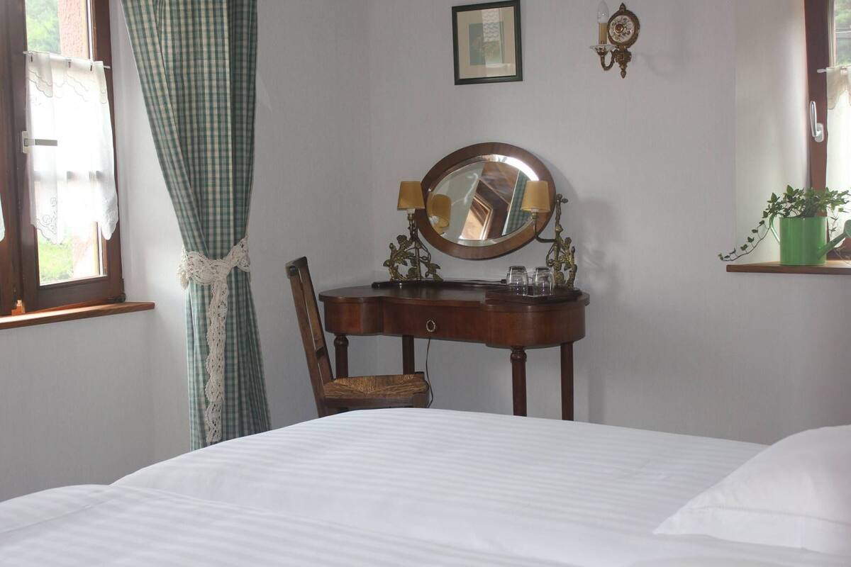 Un dormitorio con una cama, un aparador de madera con espejo, dos apliques de pared, una ventana con cortinas y una planta pequeña en un estante.