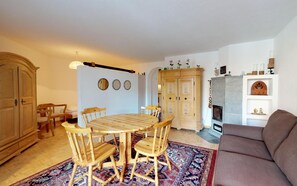 A cozy living room with a brown sofa, a wooden dining table with four chairs, a patterned rug, and a fireplace.