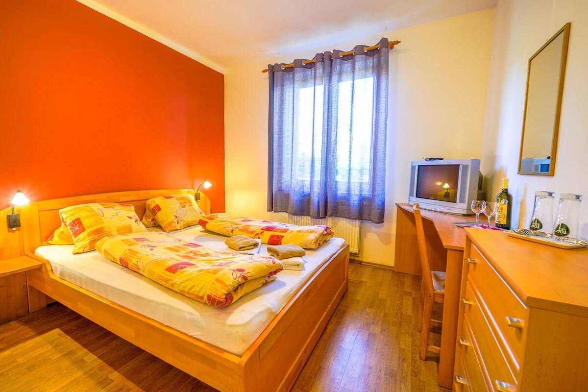 Uma cama de casal com colcha colorida, mesa de cabeceira com abajur, televisão, cômoda e espelho.