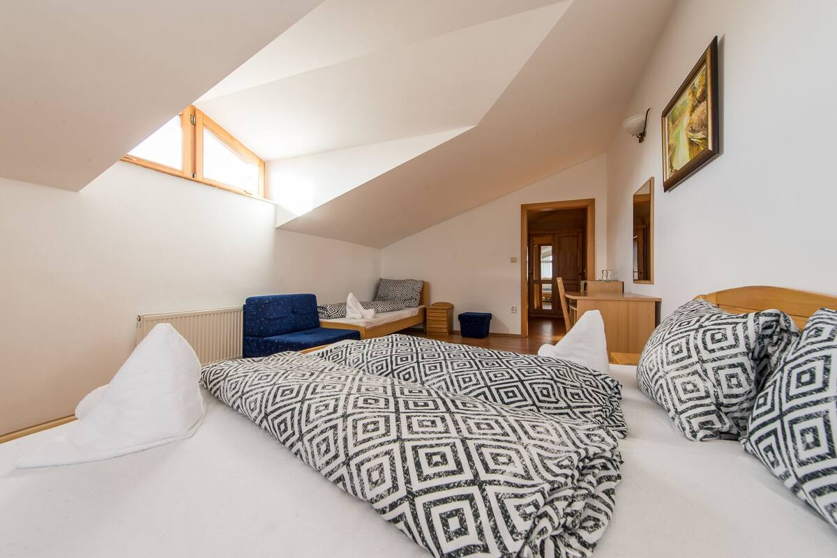 Um quarto com teto inclinado, cama com roupa de cama estampada, uma cadeira azul, mesa de cabeceira de madeira e um quadro na parede.
