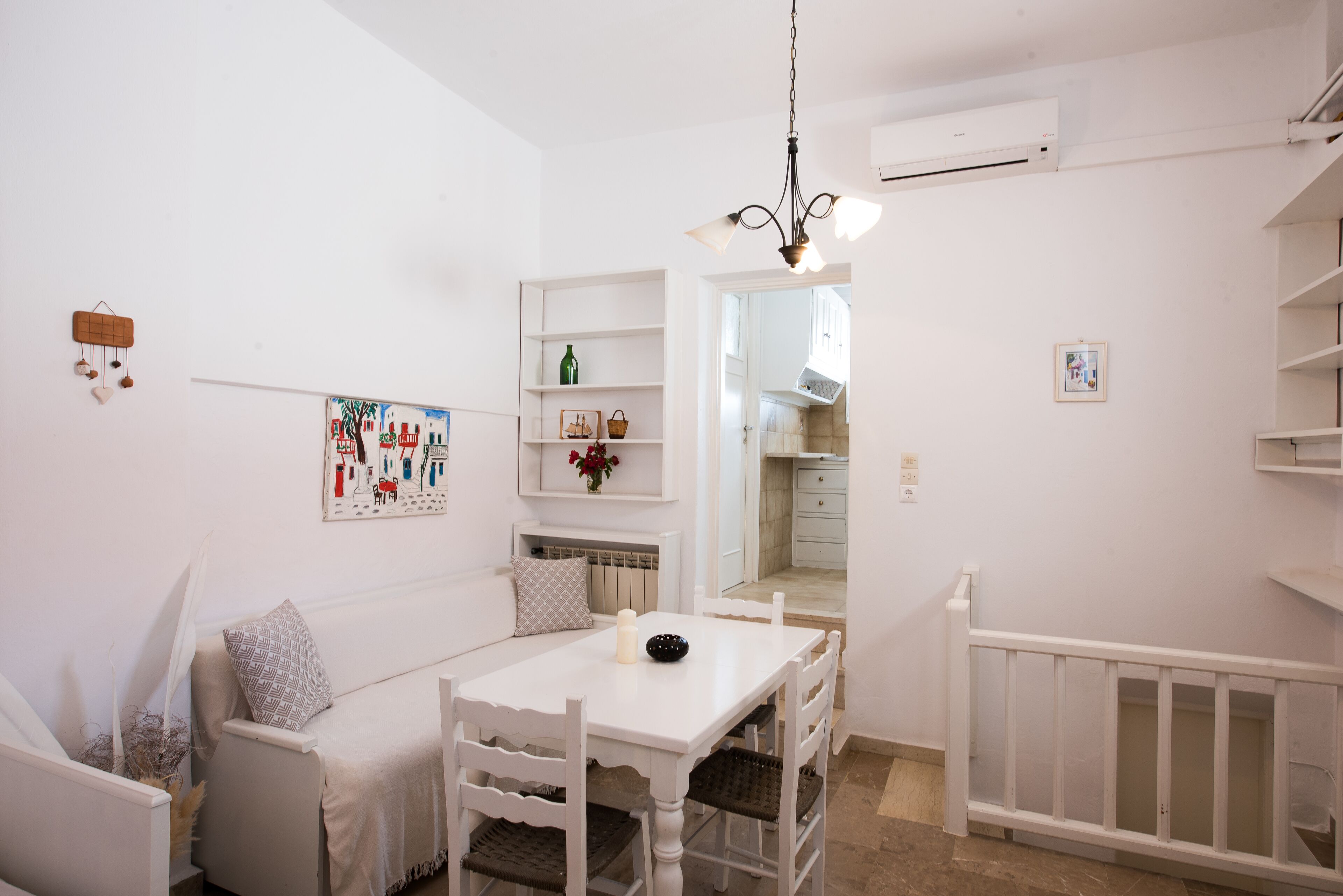 Una habitación blanca con una mesa de comedor, sillas, un banco blanco, una lámpara colgante, un estante montado en la pared con objetos decorativos, y una puerta que da a otra habitación.