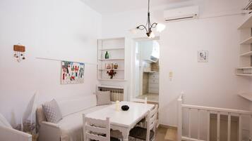 Una habitación blanca con una mesa de comedor, sillas, un banco blanco, una lámpara colgante, un estante montado en la pared con objetos decorativos, y una puerta que da a otra habitación.