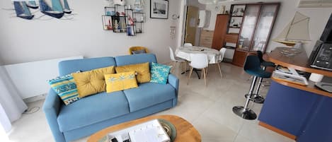 A living room with a blue sofa, a wooden coffee table, a dining area with a white table and chairs, and a kitchenette with a blue countertop.