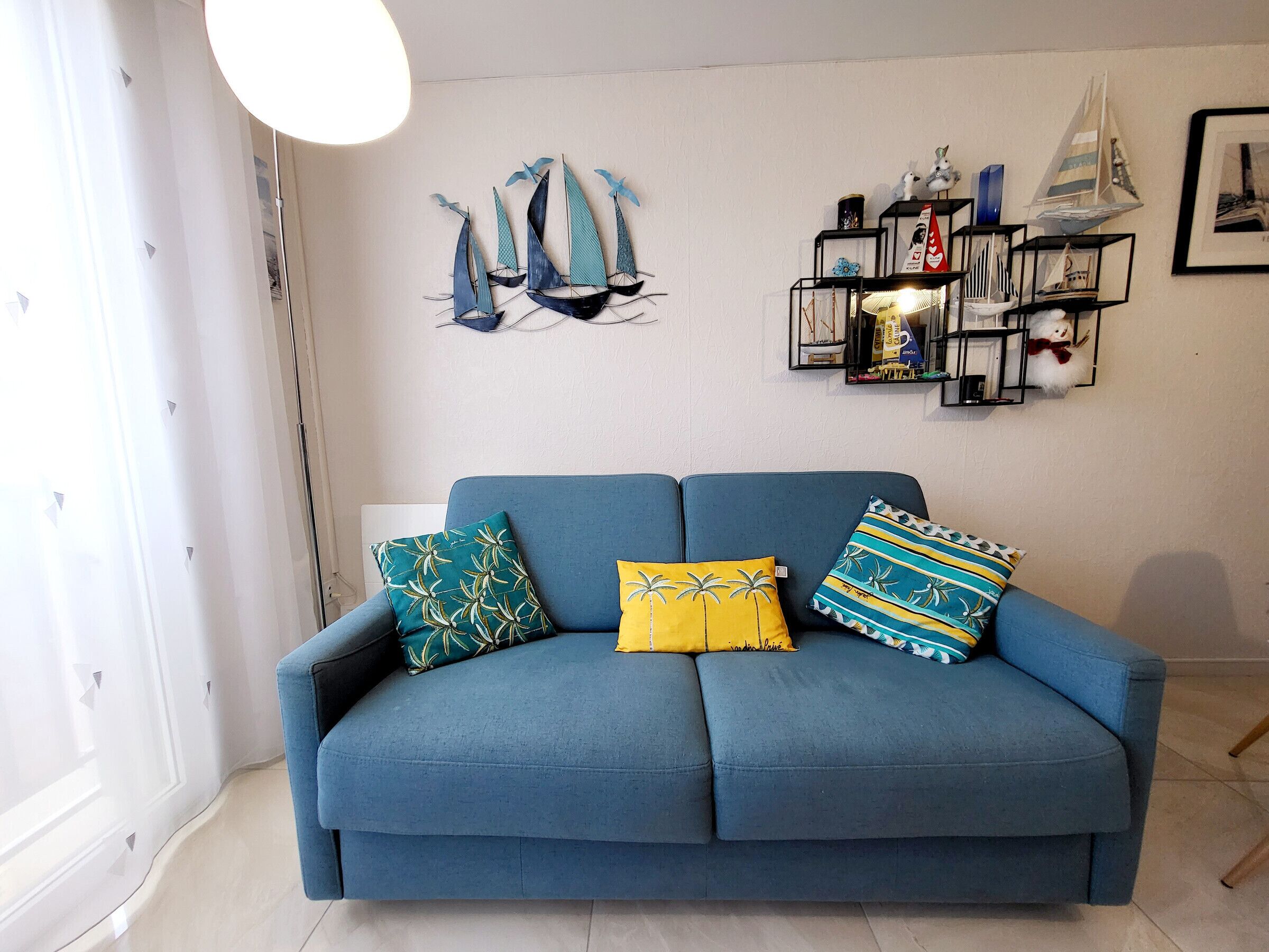 A blue sofa with three patterned cushions, a wall-mounted shelf with decorative items, and a window with sheer curtains.