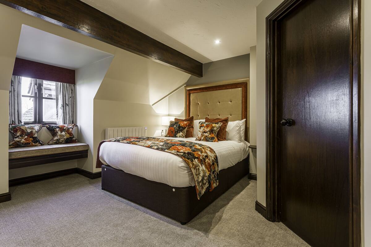 Un dormitorio con una cama grande, un asiento junto a la ventana, una puerta de madera oscura y un techo inclinado.