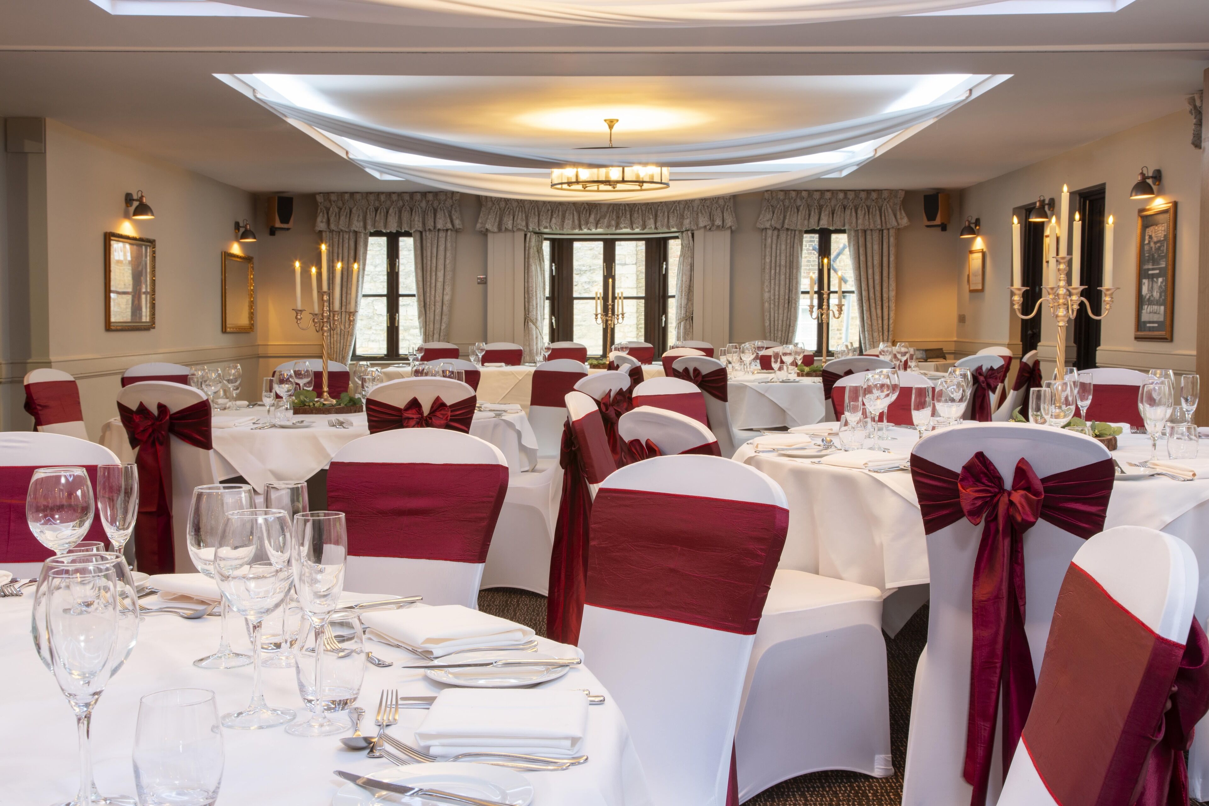 Un salón de banquetes con mesas redondas dispuestas para el servicio de comidas, cada una con manteles blancos y fajas rojas.