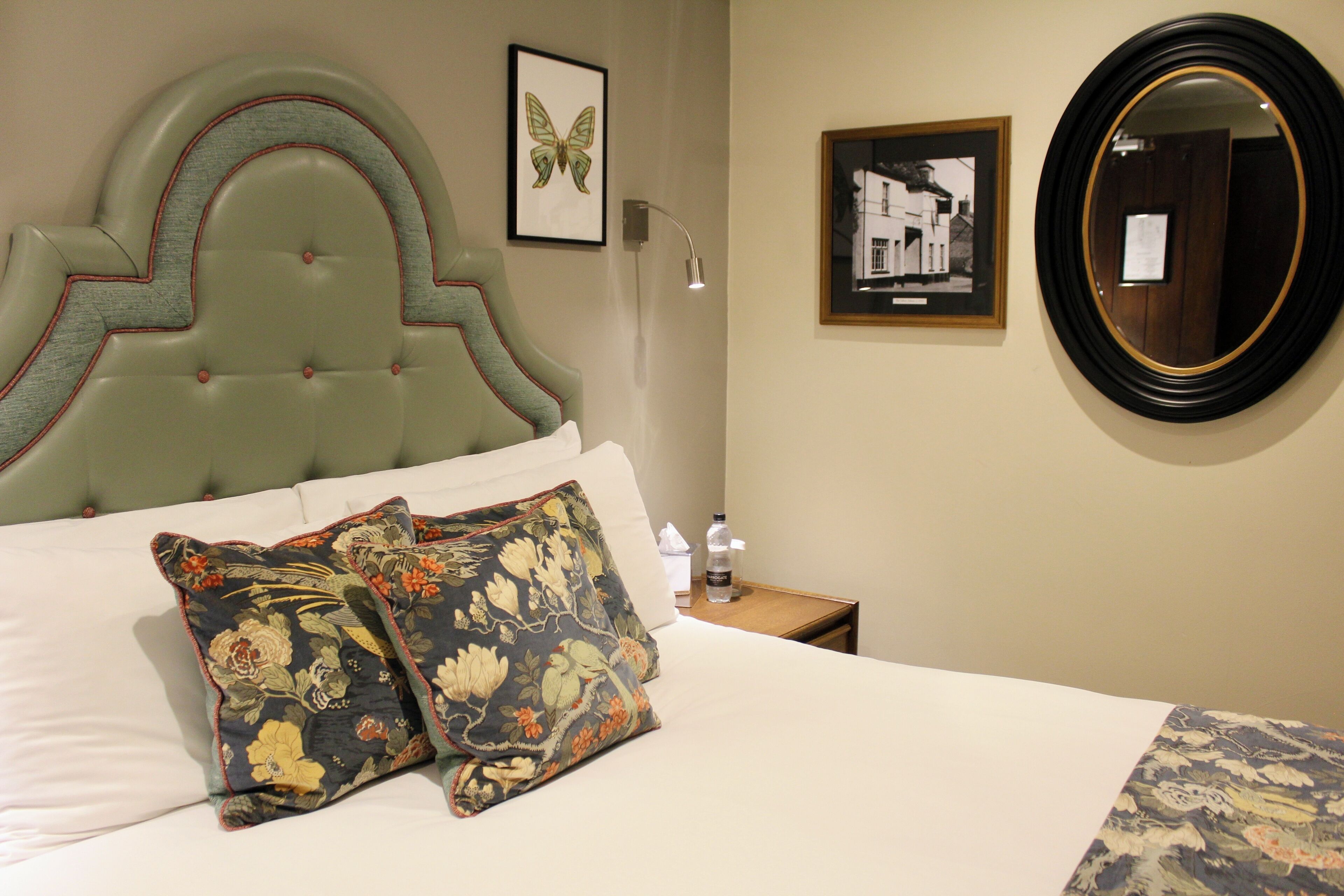 A neatly made bed with floral pillows, a framed butterfly artwork, a framed photograph, and a round mirror on the wall.