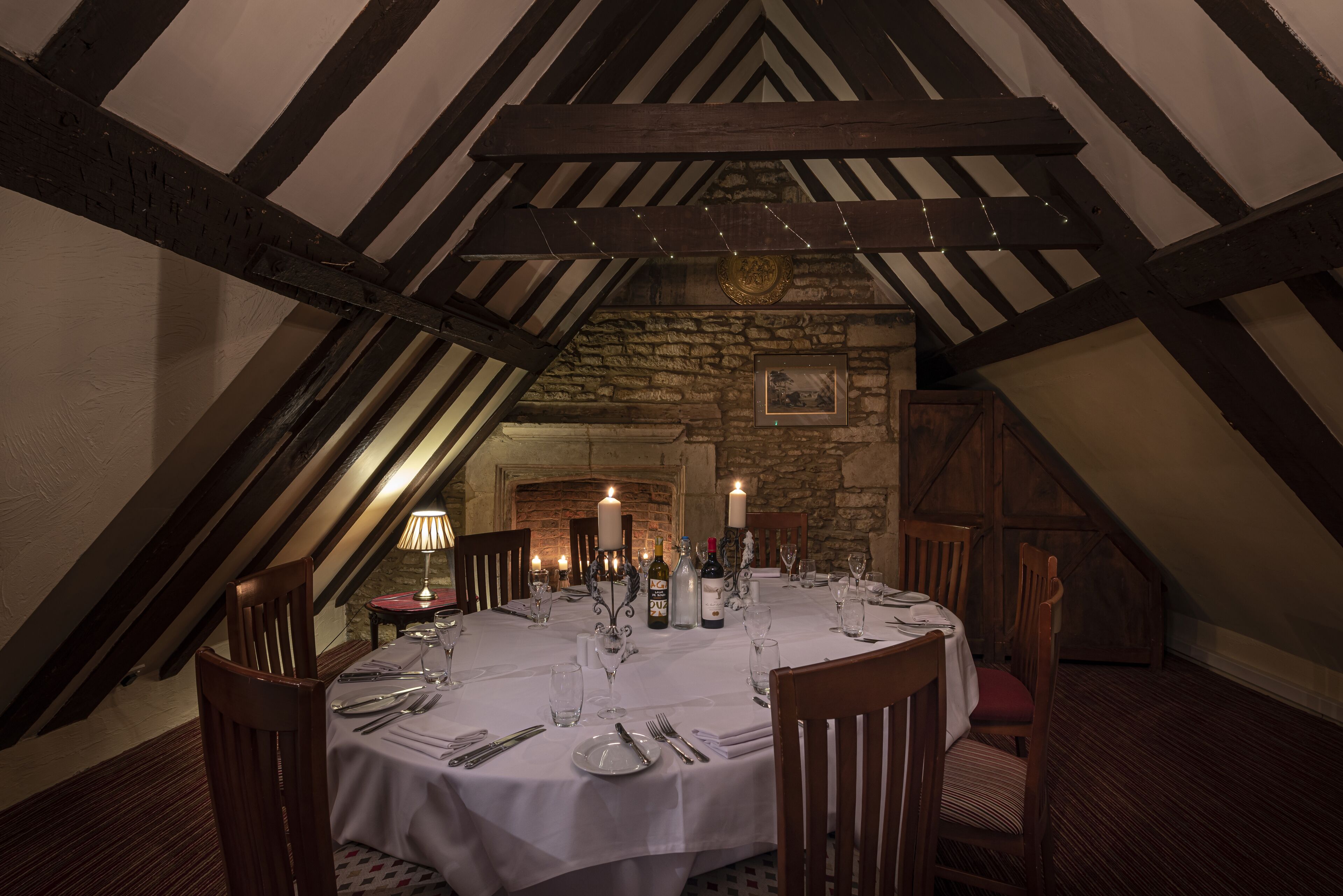 A dining room with a round table set for six, featuring white tablecloths, candles, and wine bottles. The room has exposed wooden beams and stone walls.