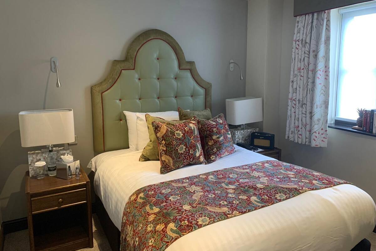 Una cama bien hecha con una colcha estampada, una mesita de noche de madera con una lámpara y una ventana con cortinas.