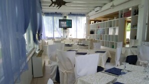 Un comedor con mesas redondas dispuestas para una comida, sillas blancas y un televisor montado en la pared.