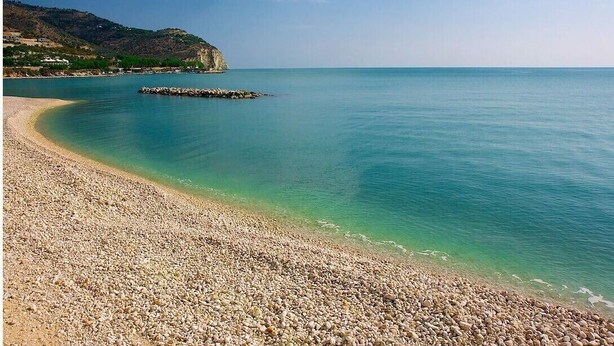 Una playa de guijarros con agua turquesa cristalina y un promontorio rocoso a lo lejos.