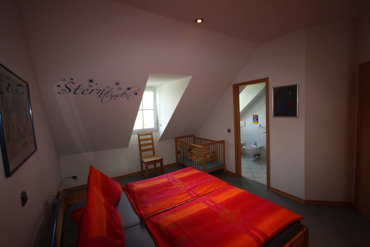 Een slaapkamer met een bed, een kinderbedje, een stoel, een raam en een zichtbare badkamer door een open deur.
