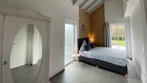 Ein Schlafzimmer mit einem großen Bett, einer Holzbalkendecke, einem kleinen Nachttisch und Blick ins Freie durch ein Fenster.