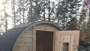 Eine kleine Holzhütte mit offener Tür, durch die ein Innenraum mit einer Bank und einem Fenster erkennbar ist.