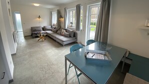 Ein modernes Wohnzimmer mit einem grauen Sofa, einem Glastisch und Blick ins Freie durch große Fenster.