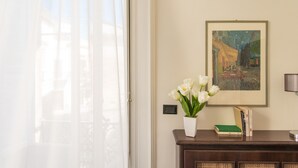 Una habitación con un amplio ventanal, un cuadro enmarcado en la pared, un florero blanco con flores y un aparador de madera con libros.