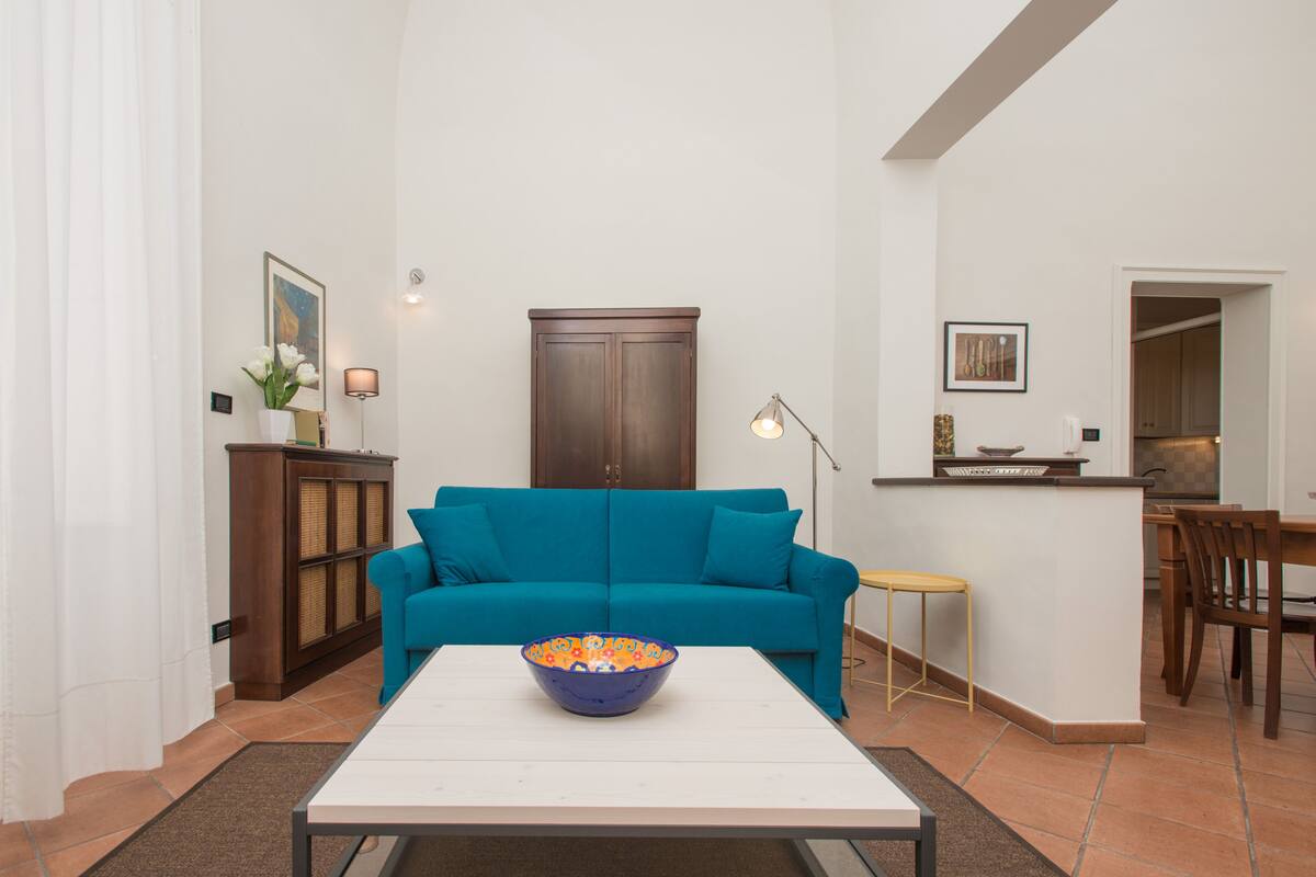 A living room with a teal sofa, a white coffee table, and a kitchen area in the background.