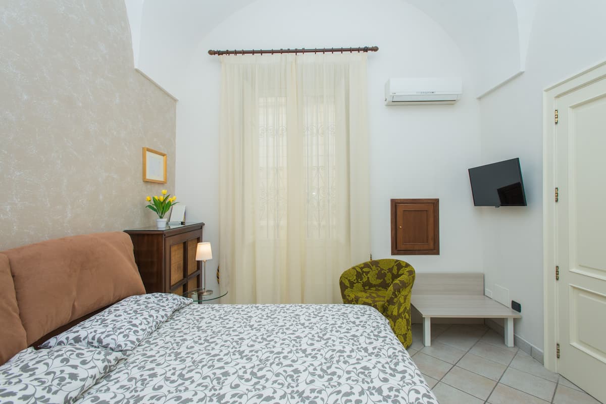A bedroom with a bed, a bedside table with a lamp, a chair, a TV mounted on the wall, and a door.