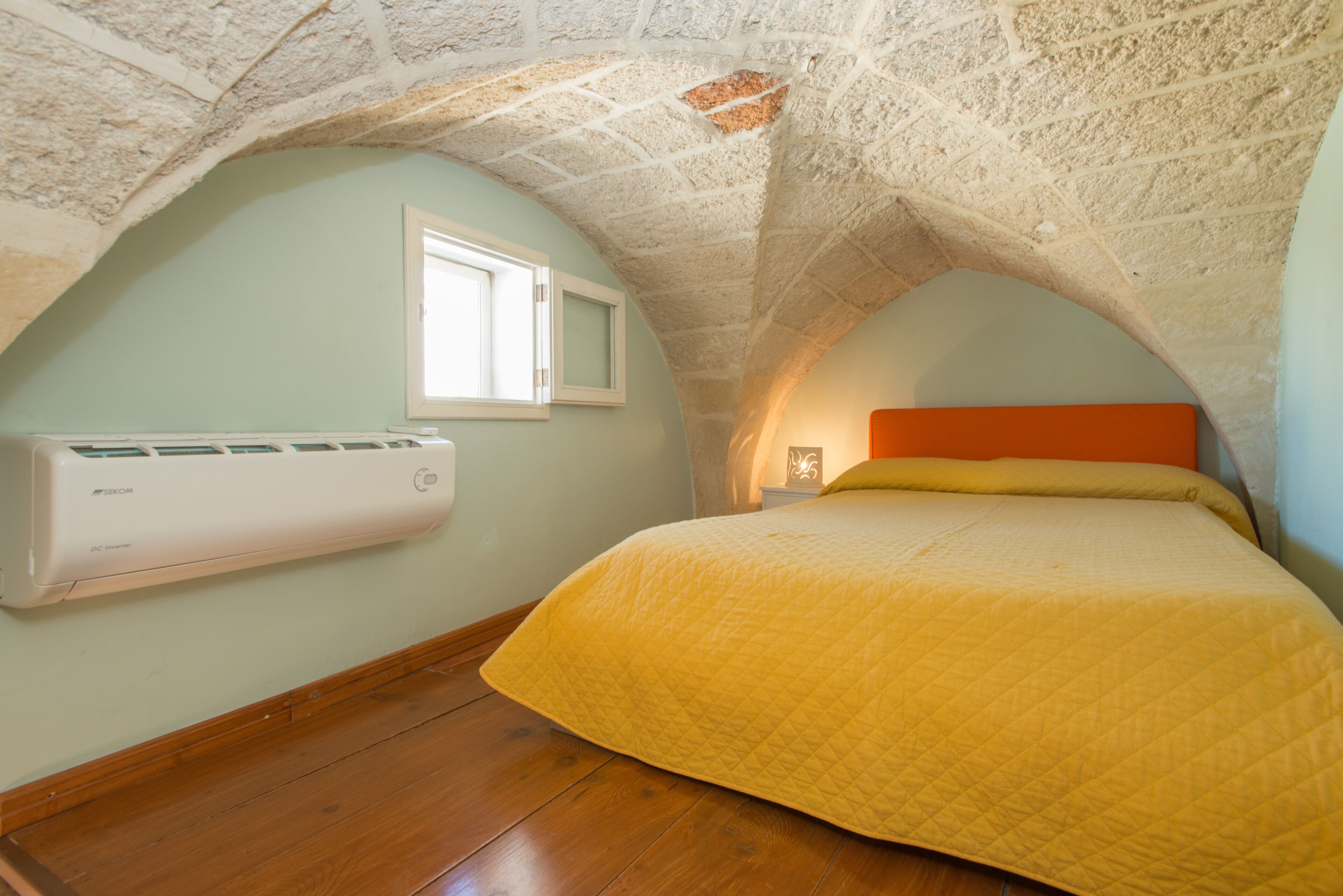 A bedroom with a sloped ceiling, a bed with a yellow quilt, a window, and an air conditioning unit.