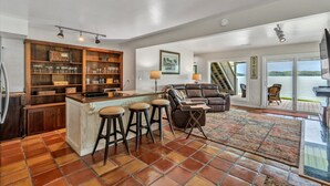 A spacious living area with a kitchen island, bar stools, a leather sofa, and a large window overlooking a scenic view.