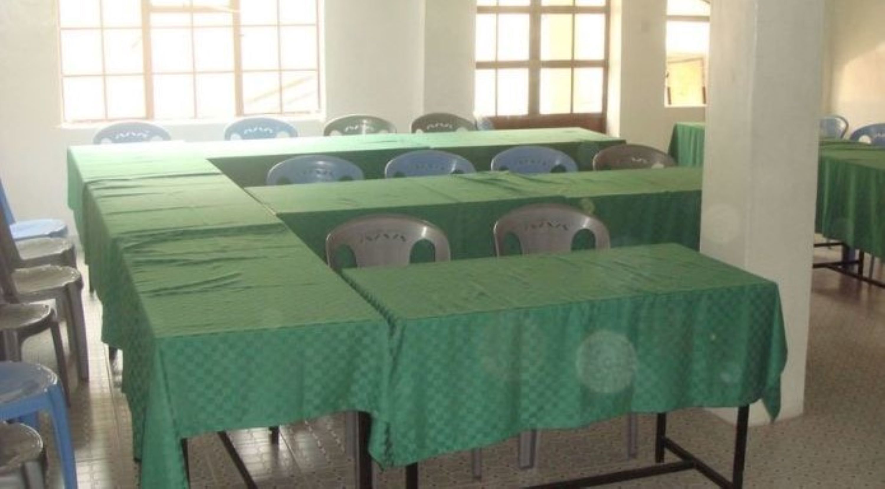 Un salón de conferencias con mesas y sillas verdes, ventanas amplias y suelo de baldosas.