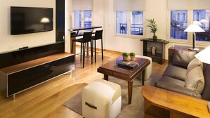 A modern living room with a flat-screen TV, a dining area with chairs, a sofa, a coffee table, and a view of the city through large windows.