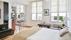 A modern hotel room with a large bed, a kitchenette, a sitting area with a table and chairs, and large windows with a view.