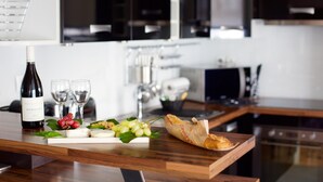 A wooden table with a bottle of wine, glasses, a cheese board with grapes, and a baguette in a modern kitchen.
