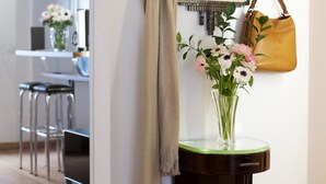 A hallway with a mirror, a vase with flowers, a hanging bag, and a bench.