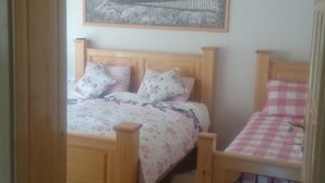 A bedroom with two wooden beds, one with a floral bedspread and the other with a checkered bedspread. There is a framed picture on the wall above the beds, a wooden ceiling fan, and a wooden door frame visible in the foreground. - Apartments Una Riverlodge (Bihac)