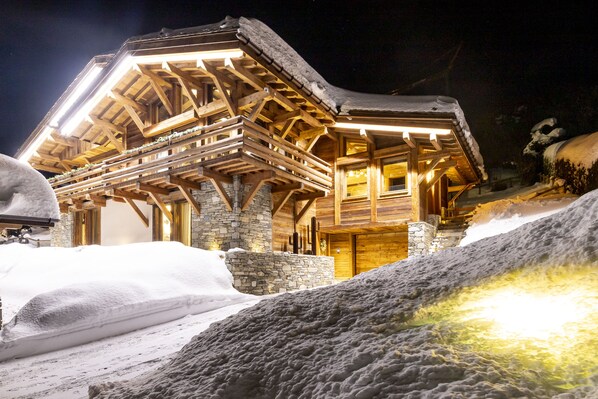 A wooden chalet with a stone foundation, wooden balconies, and a lit entrance. - CHALET GRAND PARADIS, comfort, refinement, 7 bedrooms, cinema, SPA, near center (Megève)