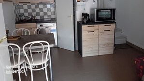 Cozinha compacta com armários de madeira, micro-ondas e uma televisão pequena. Há uma área de jantar com mesa e cadeiras.