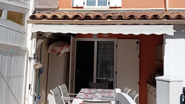 Um imóvel residencial com área de varanda equipada com mesa e cadeiras, porta deslizante e pequena cozinha visível por uma porta aberta.