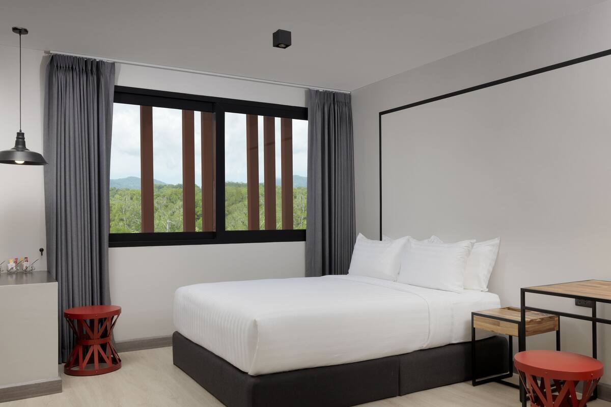 A modern bedroom with a large bed, a small table, two red stools, a pendant light, and a window with curtains.
