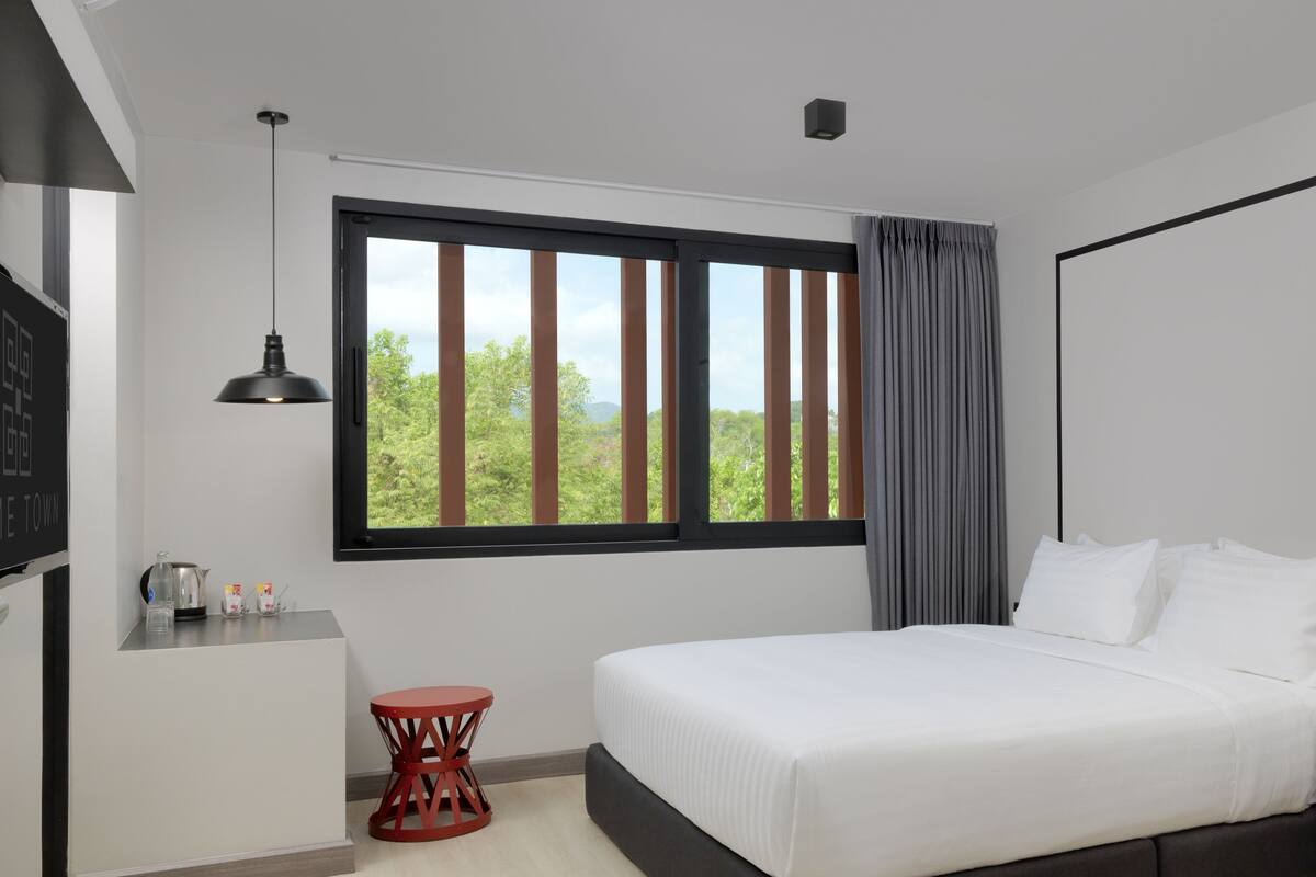 A modern hotel room with a large bed, a small table, a red chair, and a window with a view of greenery.