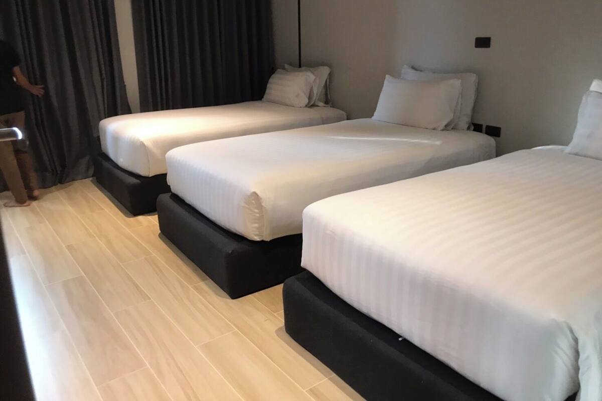 A hotel room with three single beds, a wooden floor, and a dark curtain.