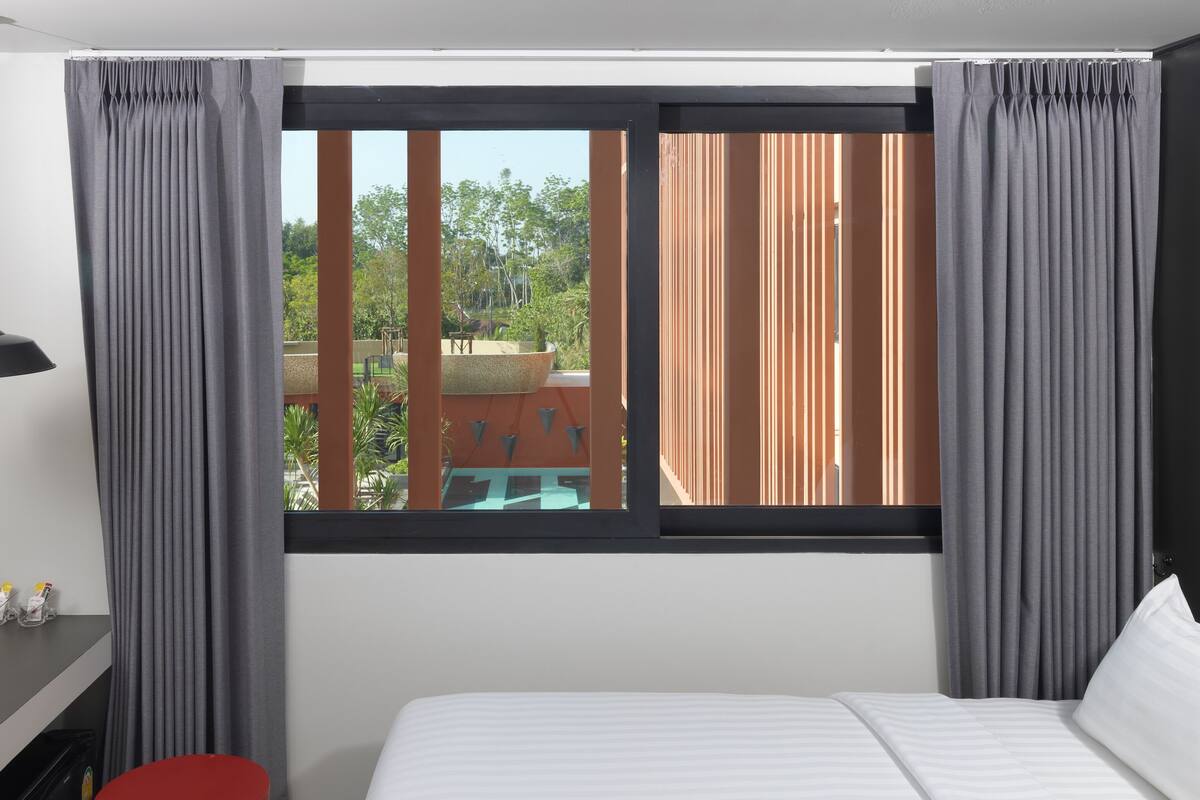 A hotel room with a bed, a desk with a kettle, cups, and bottles, and a window with curtains.