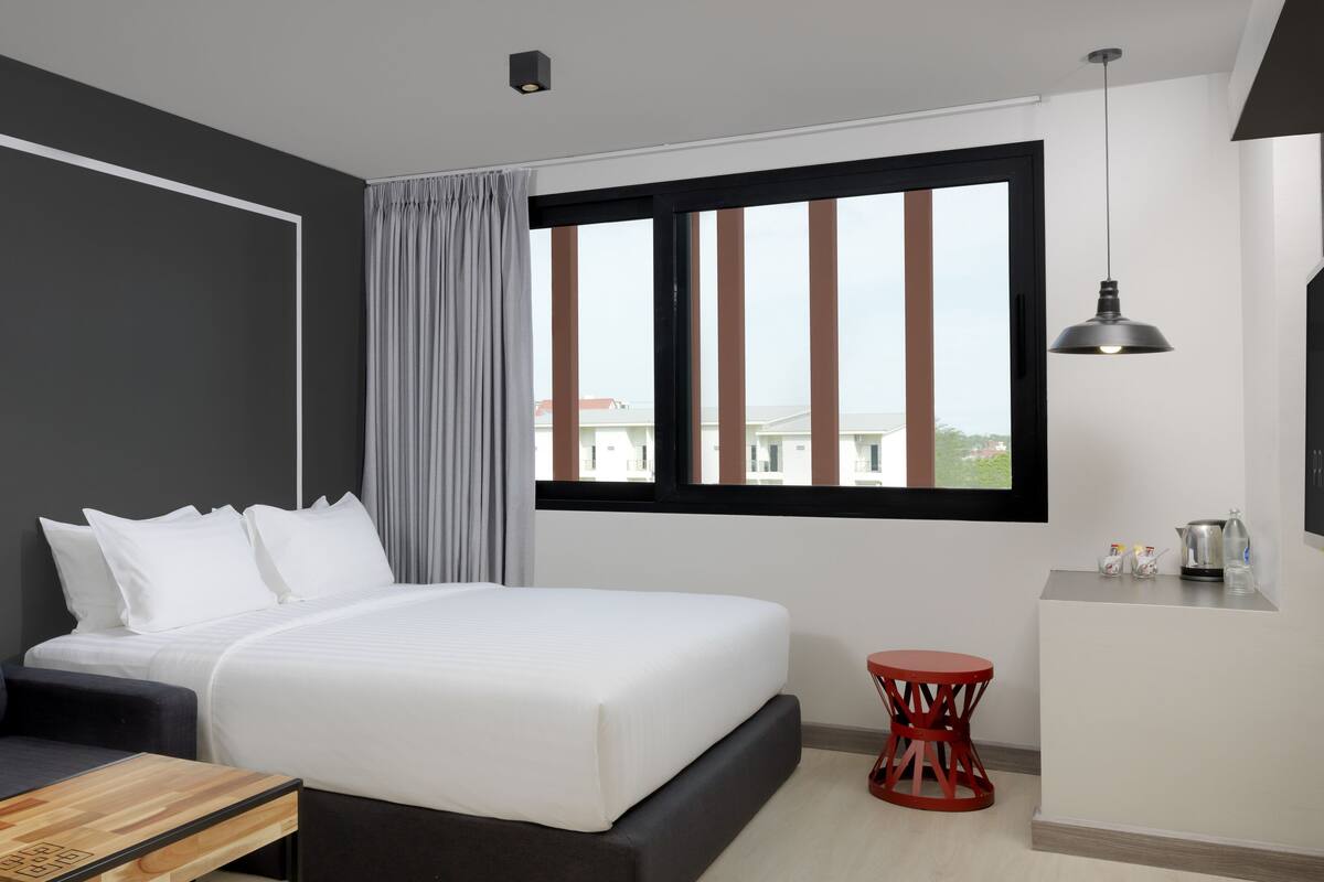 A modern hotel room with a large bed, a sofa, a small table, a red chair, a window with curtains, and a view of buildings outside.