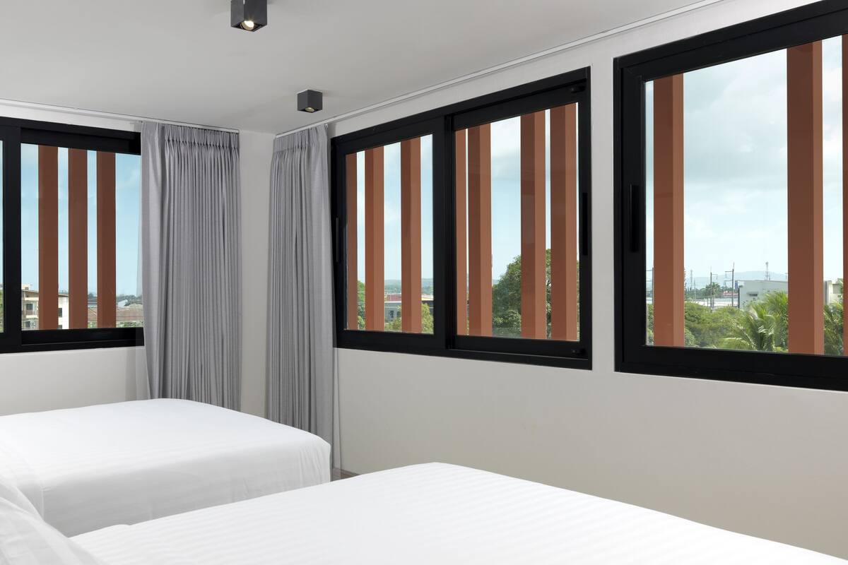A hotel room with two beds, large windows with vertical blinds, and a view of the cityscape.