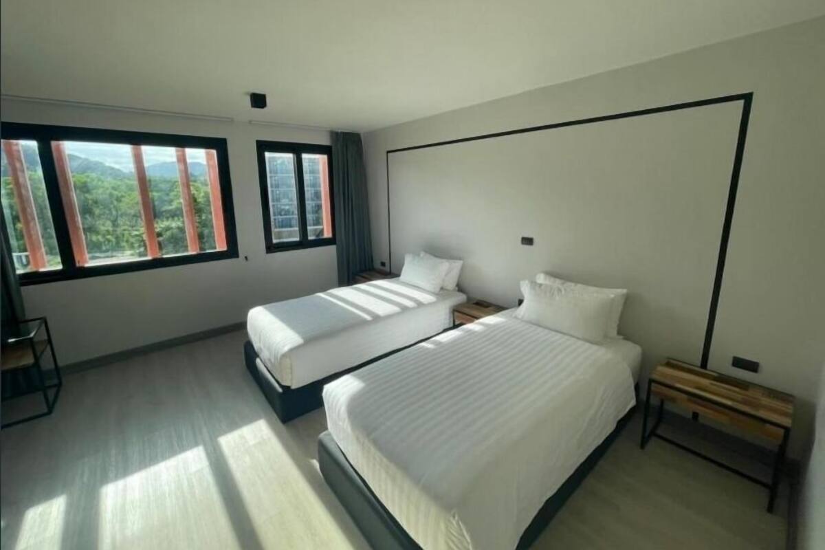 A modern hotel room with two beds, large windows with curtains, a wooden bench, and a minimalist design.