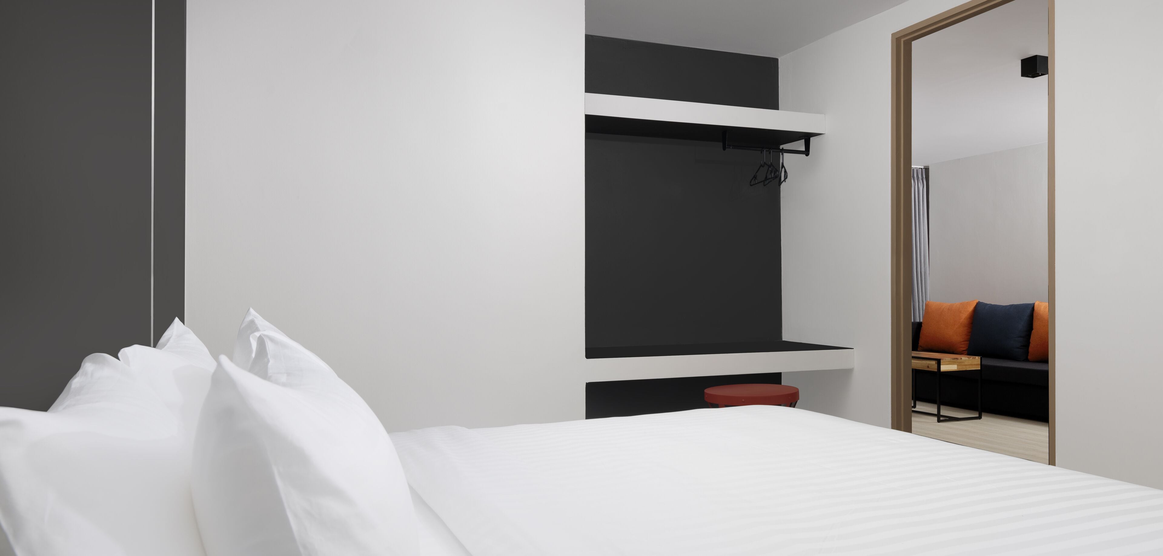 Et moderne hotelværelse med en seng, en rød skammel, en sort hylde og et spejl, der afspejler en sofa og et bord.