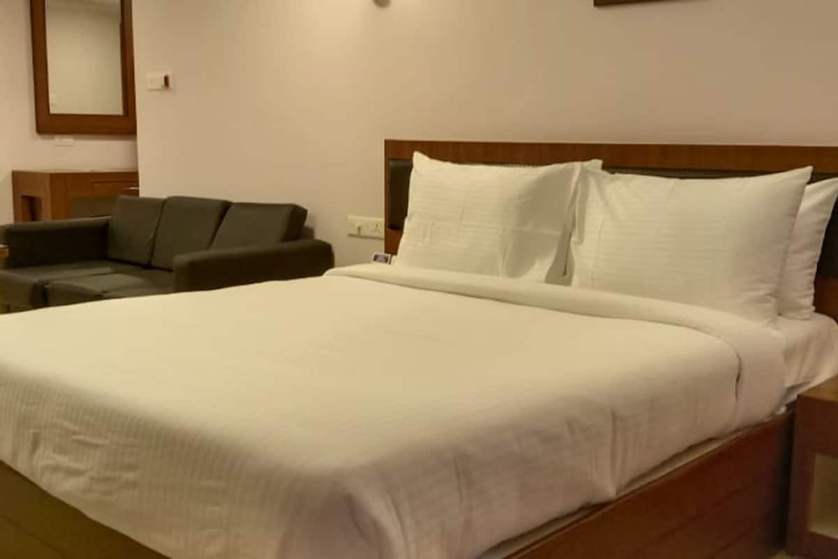 A hotel room with a large bed, a wooden headboard, a nightstand, a bedside table, and a dark-colored sofa.